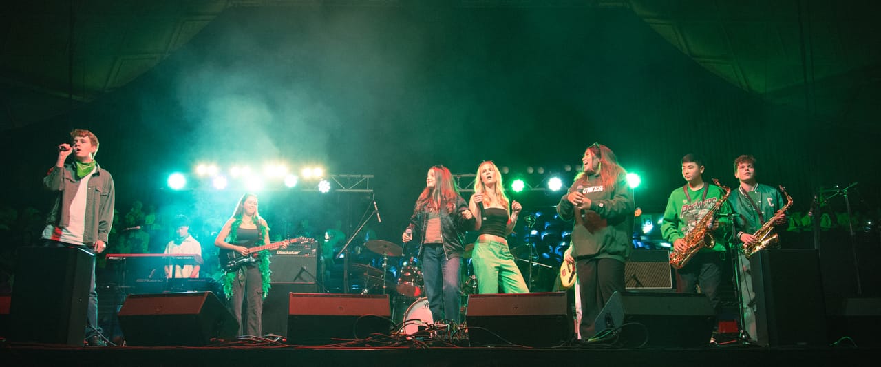 Performers with horns and guitars on stage under green and white lights.