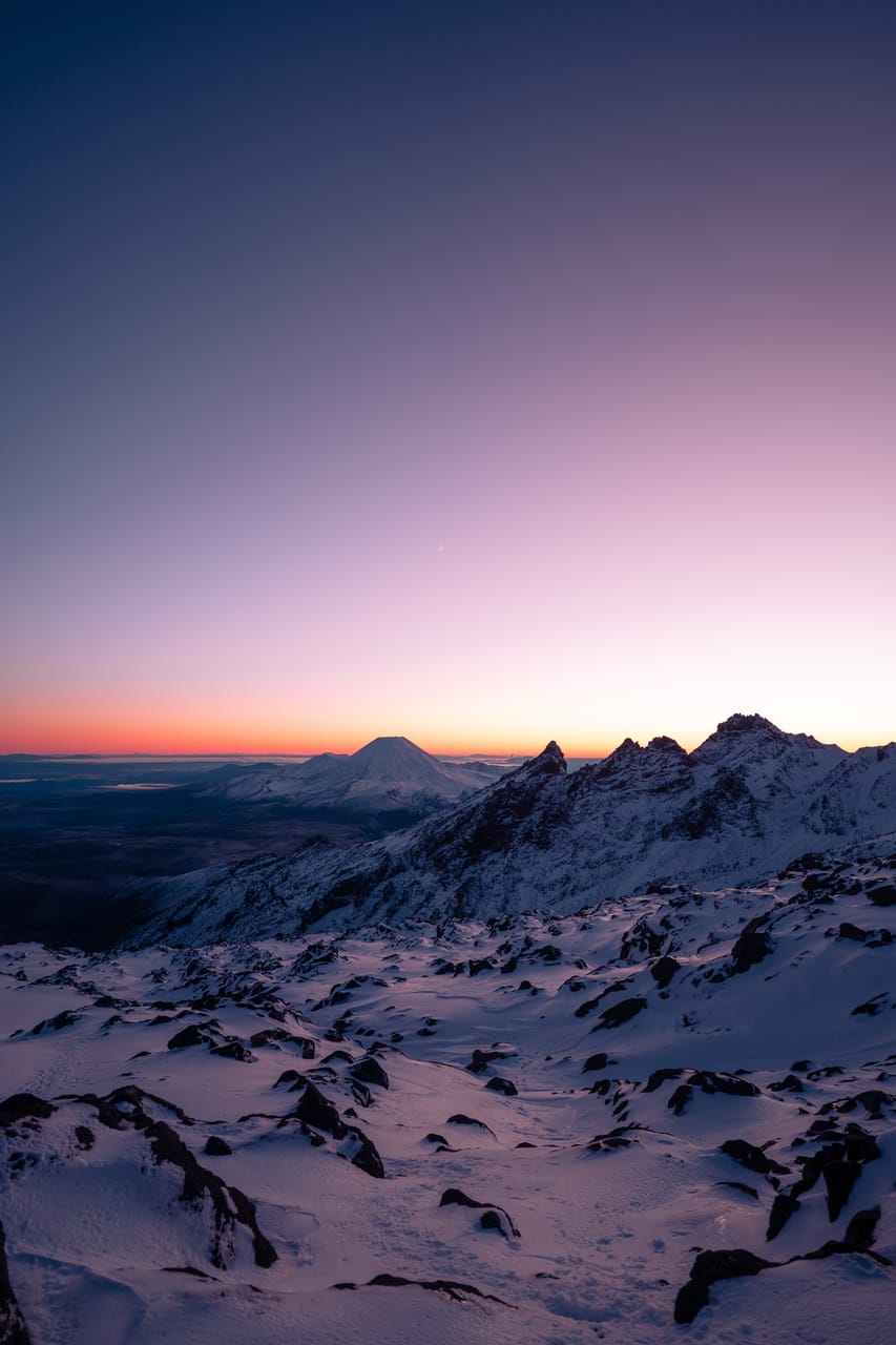 Snowy alpine ridges under a pink twilight sky, with a distant volcano on the horizon.
