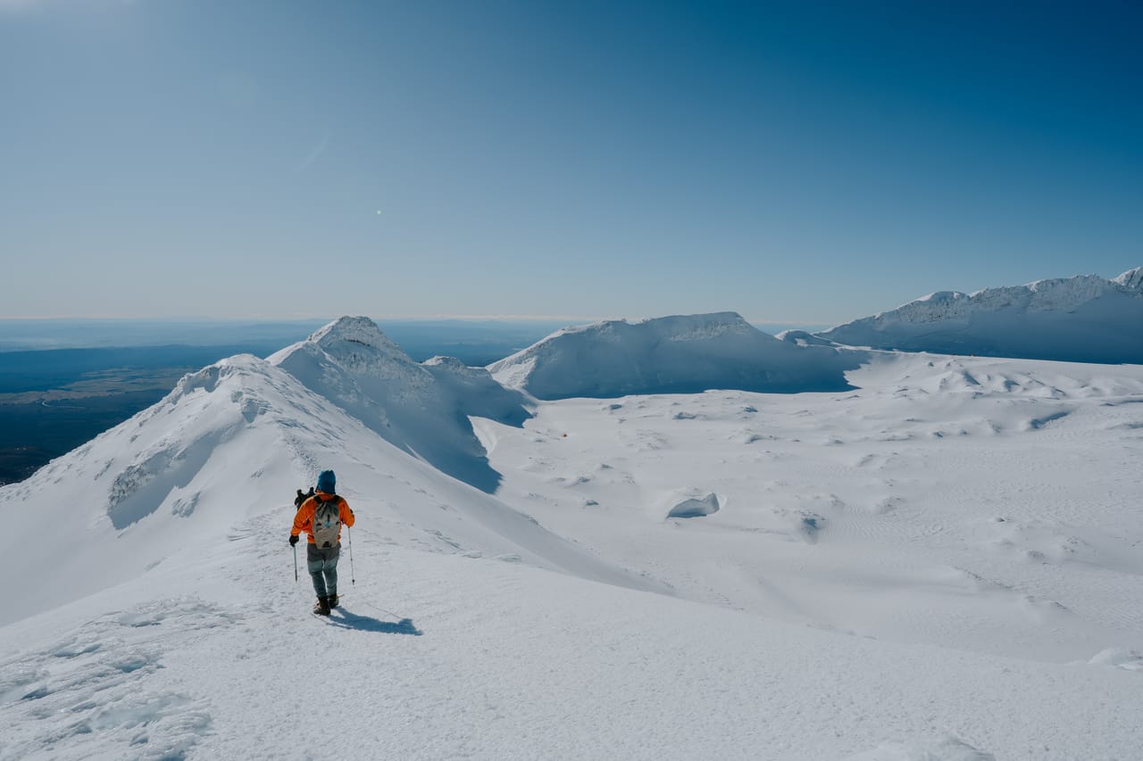 A climber in an orange jacket walks across a snowy summit plateau.