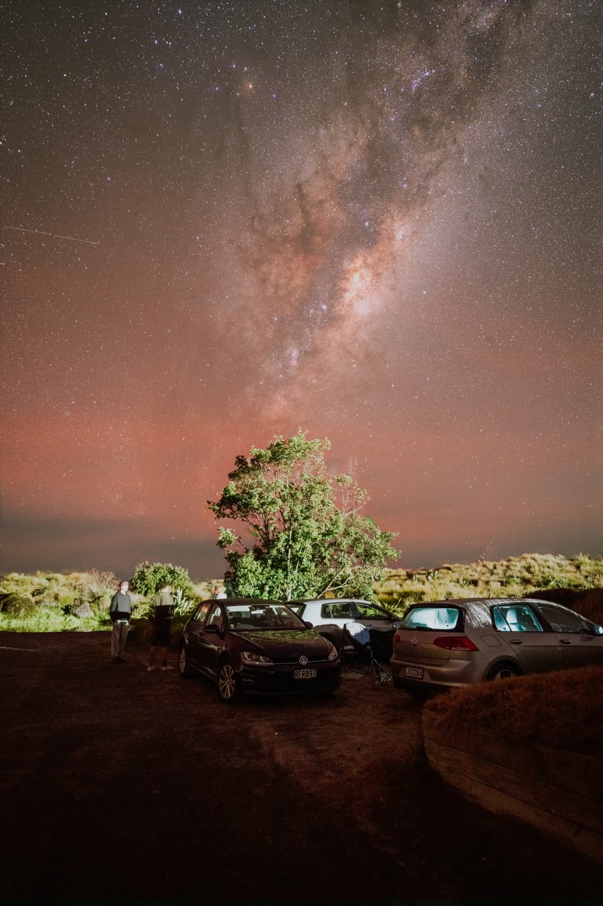 The Milky Way rises above a tree and parked cars.