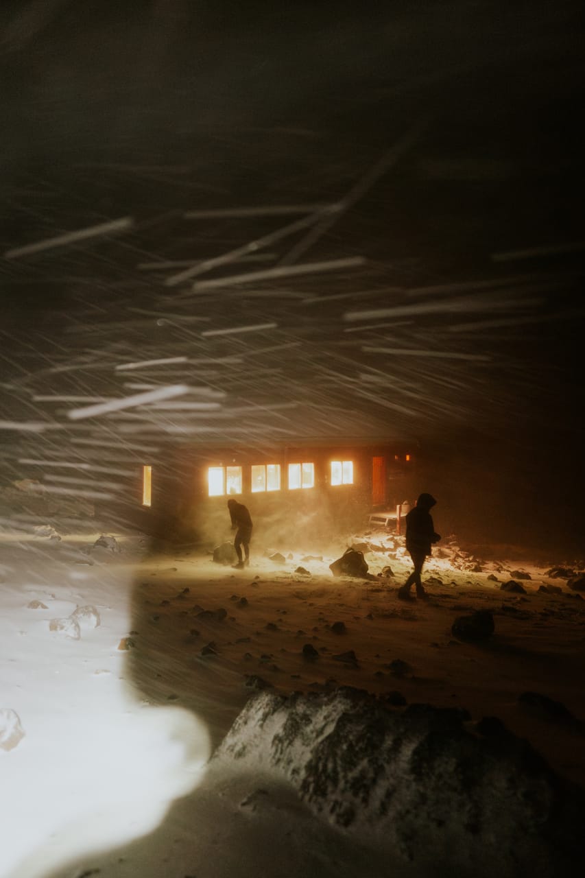 Two people outside a lit alpine hut during a night snowstorm.