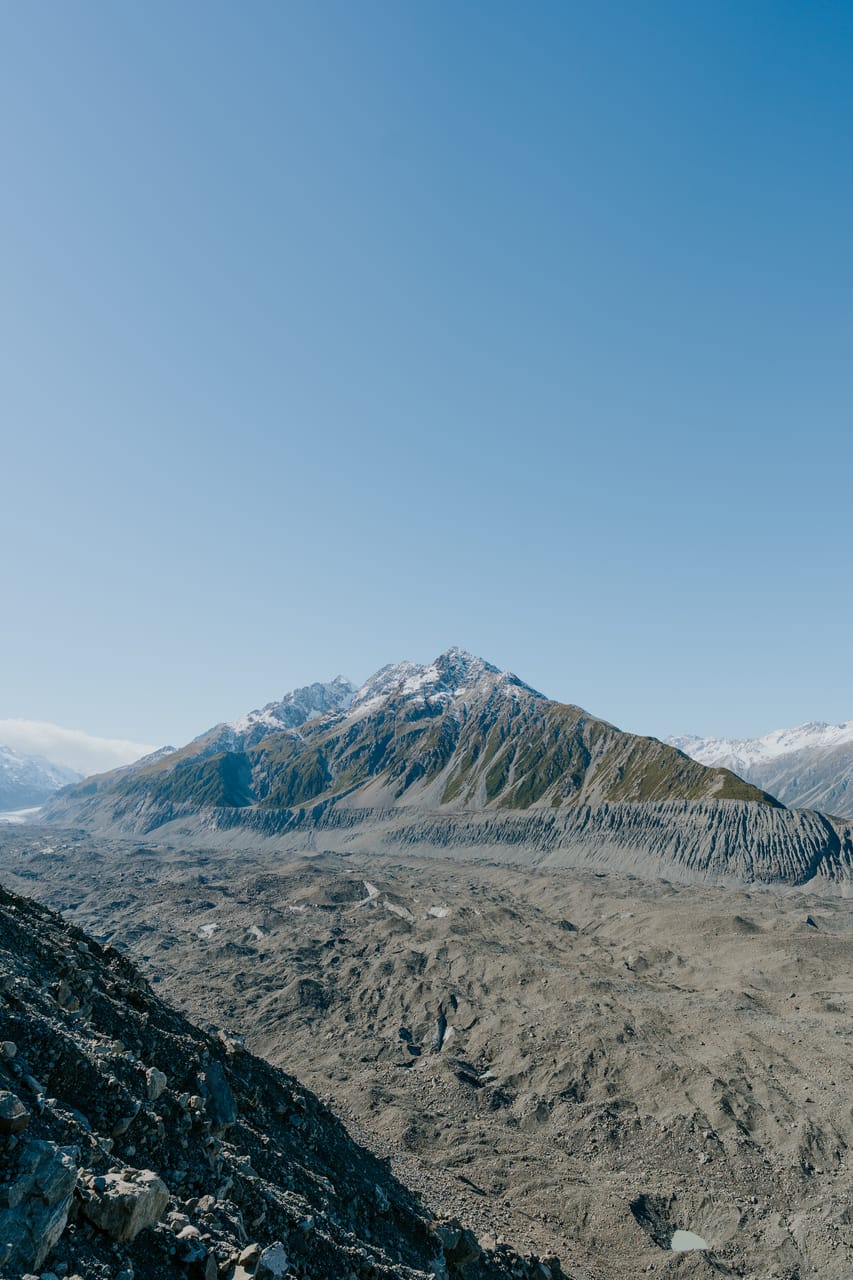 A rocky mountain rises above a glacial valley under a clear blue sky.