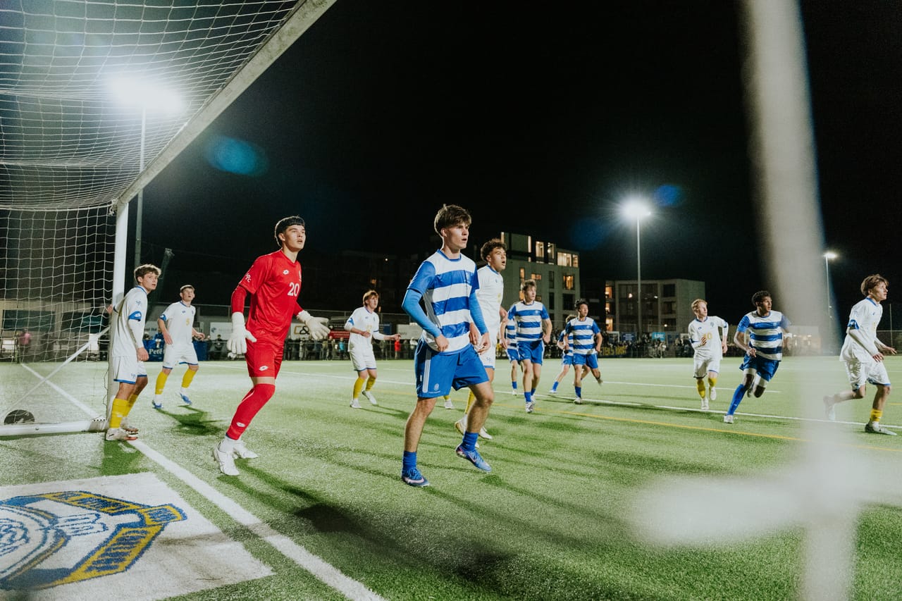 Footballers in red and in blue-and-white stripes chase the ball under floodlights.