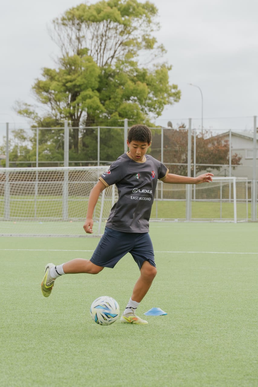 A footballer in a dark training top strikes the ball during a daytime session.