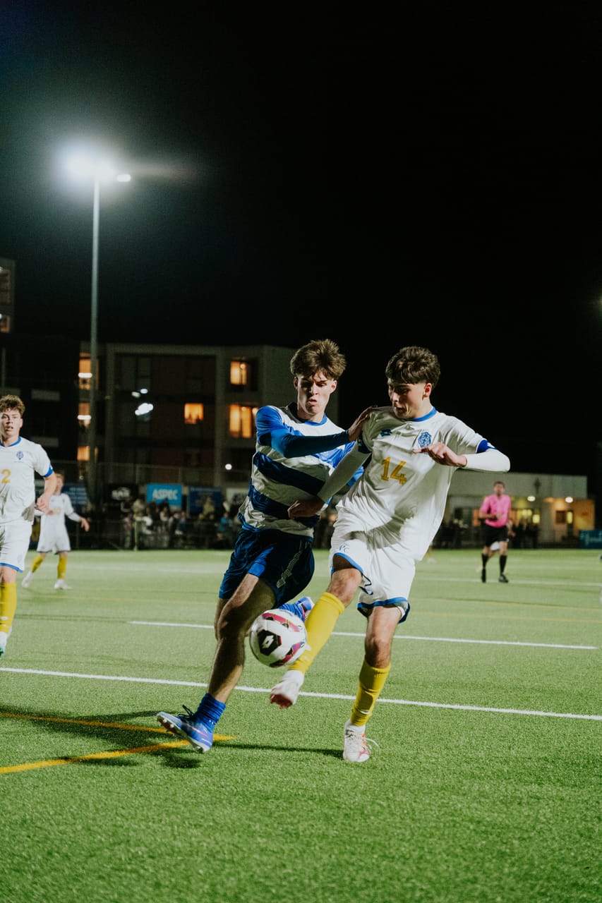 Two players battle shoulder to shoulder for the ball on a floodlit pitch.