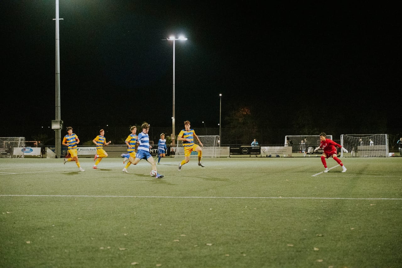 A wide view of a floodlit match as players in yellow and blue move up the pitch.