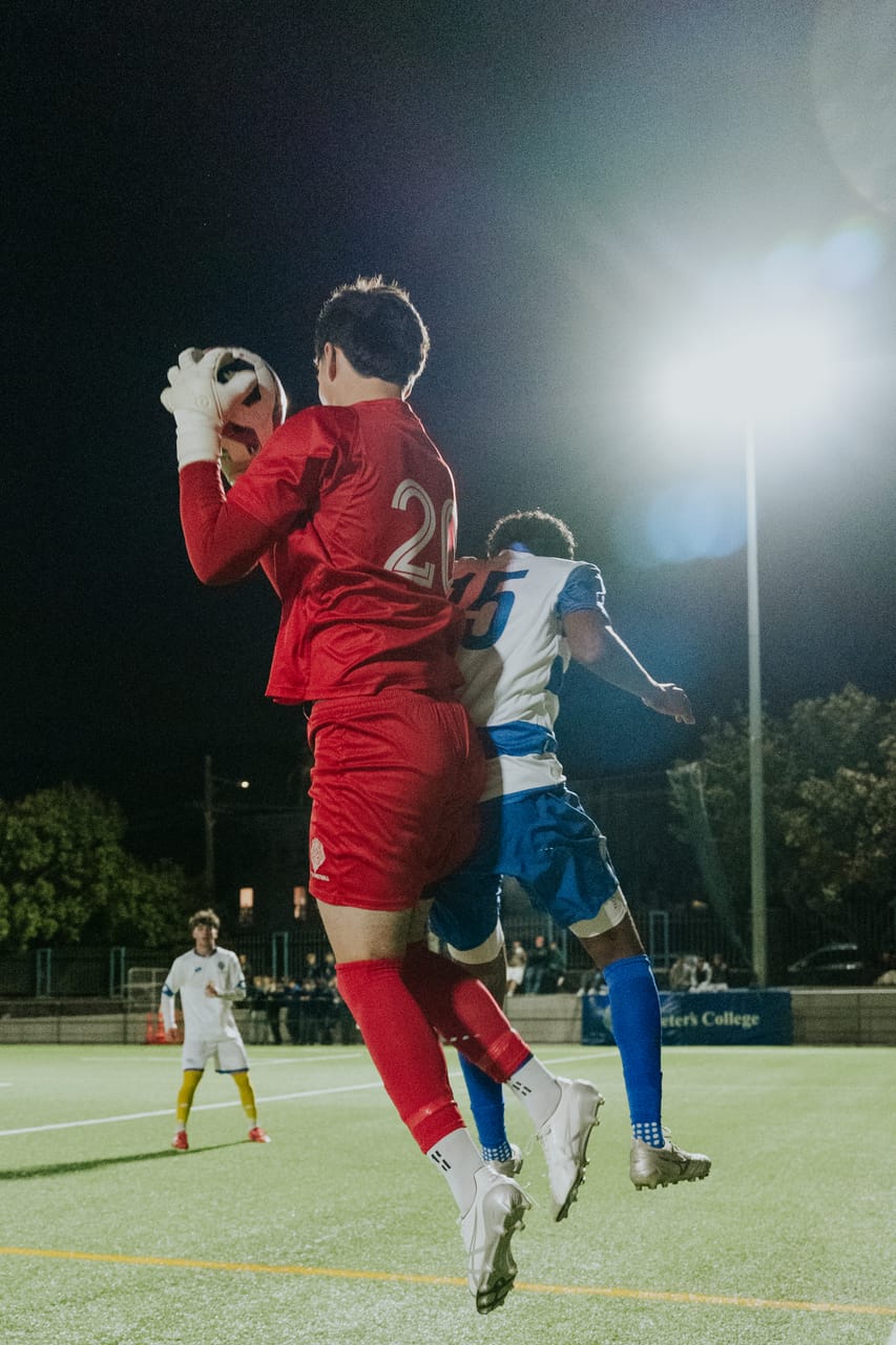 A goalkeeper in red jumps to catch a high ball under floodlights.