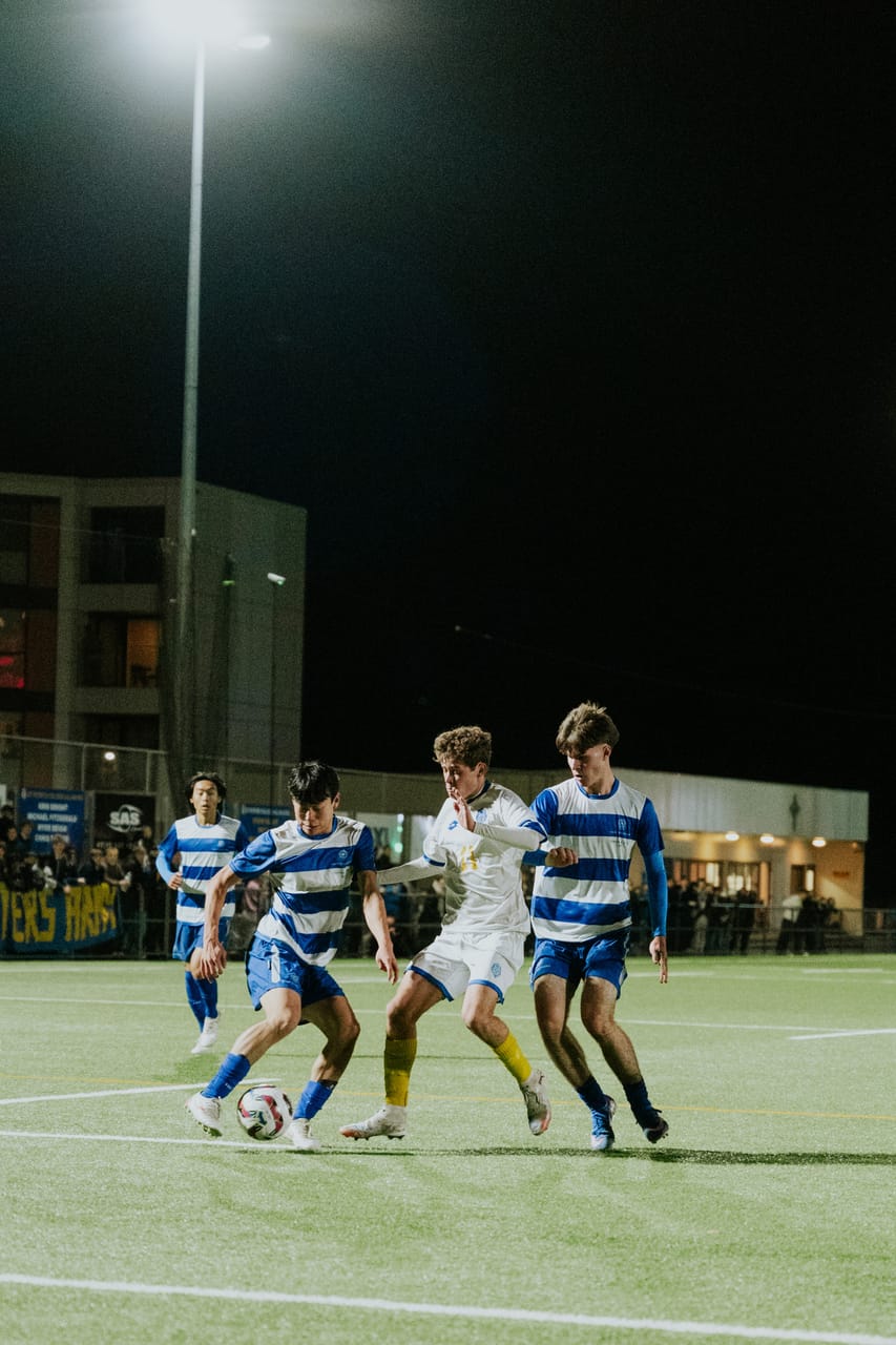 Three players in blue-and-white stripes close down a player in white on a night pitch.
