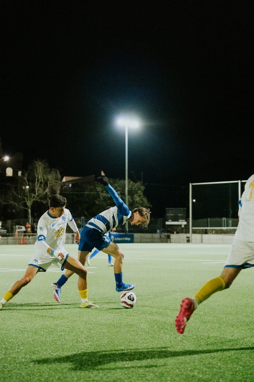 A player in blue stretches to reach the ball ahead of a player in white at night.
