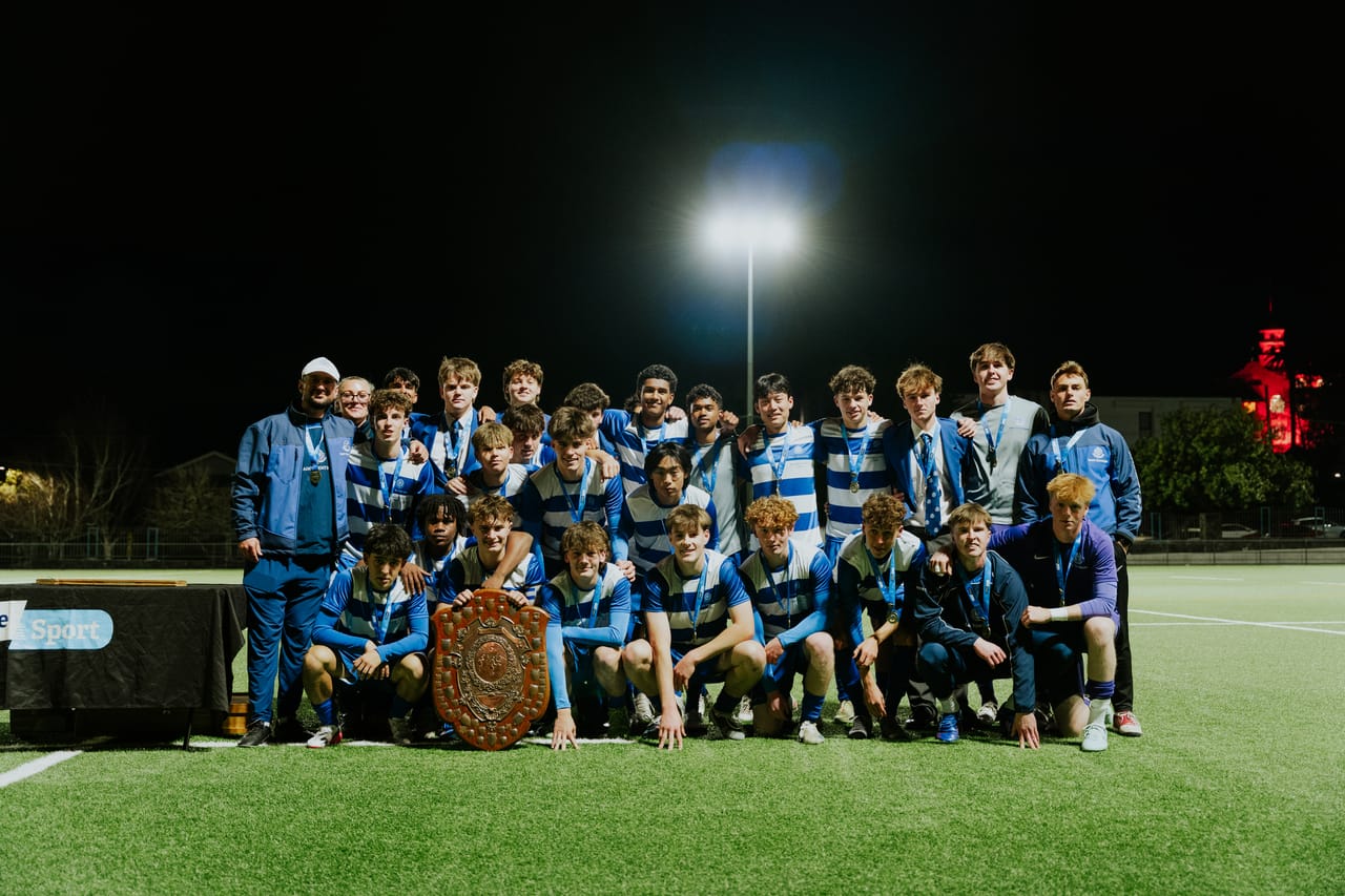 A team in blue-and-white stripes poses with a trophy shield under the floodlights.