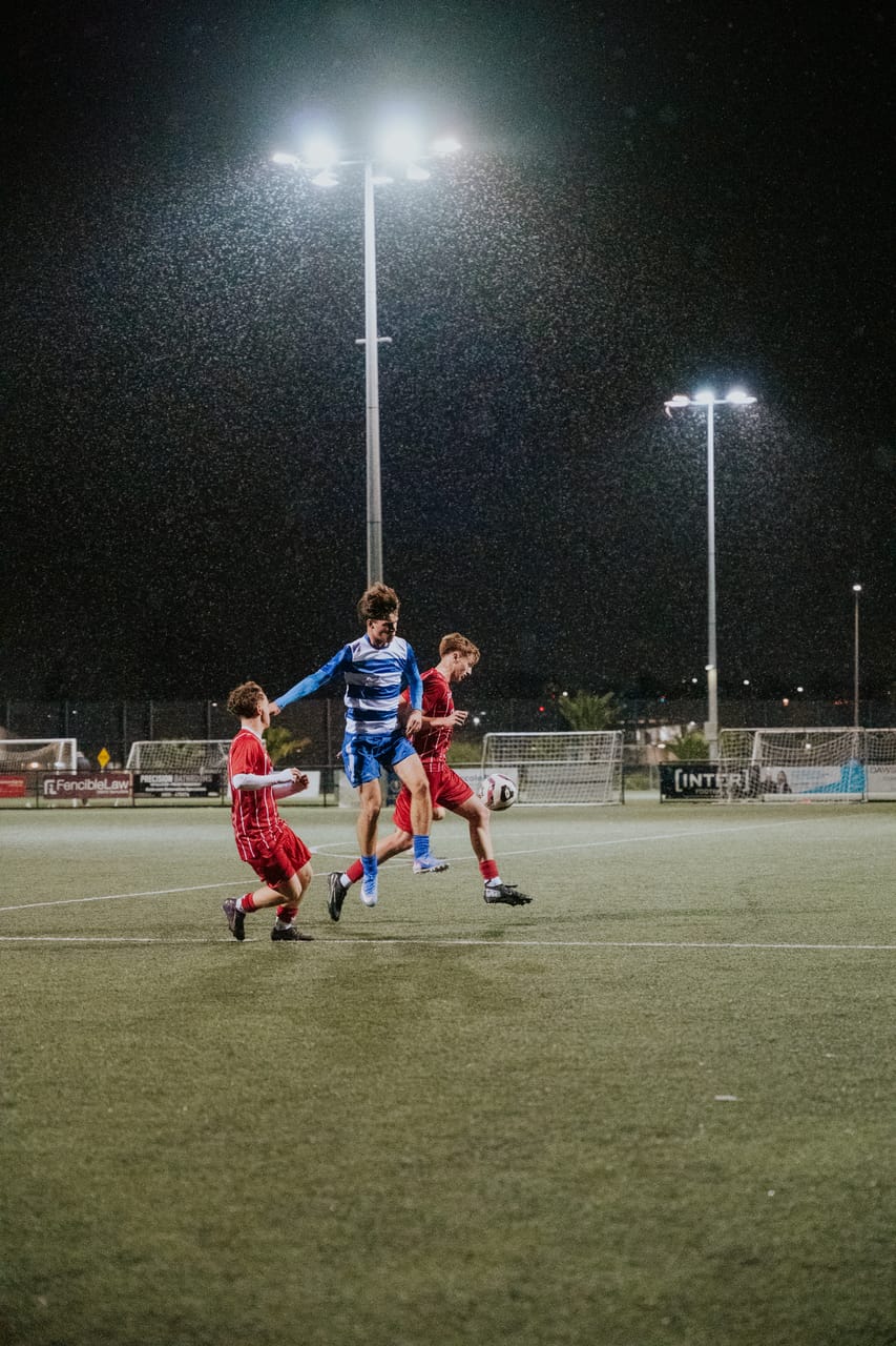 A player in blue-and-white stripes shields the ball from two players in red under floodlights.