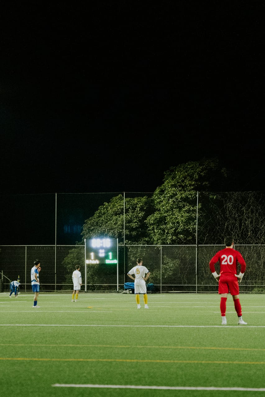 A player in red waits on a dark floodlit pitch as play continues at the far end.